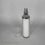 A transparent Round Airless Bottle with a metallic pump dispenser contains white lotion or cream. The transparent bottle cap is placed next to the bottle. The brand name "COMPAX" appears in the bottom left corner, and the background is plain and light-colored.