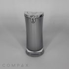 A sleek, silver Hourglass Airless Bottle with a transparent base and a metallic cap, embossed with a decorative design. The background is plain gray. The brand name "COMPAX" is in small letters at the bottom left corner.