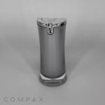 A sleek, silver Hourglass Airless Bottle with a transparent base and a metallic cap, embossed with a decorative design. The background is plain gray. The brand name "COMPAX" is in small letters at the bottom left corner.
