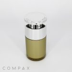 A cylindrical Round Airless Bottle of perfume with a metallic silver cap on top. The bottle is partially transparent, revealing a yellowish-green liquid inside. "COMPAX" is written in light grey at the bottom left corner of the image.
