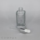 A clear, empty 30mL Glass Square Bottle with a silver cap and rubber bulb is lying on its side with the dropper separate from the bottle. The background is plain and light-colored. The word "COMPAX" is visible at the bottom left corner of the image.