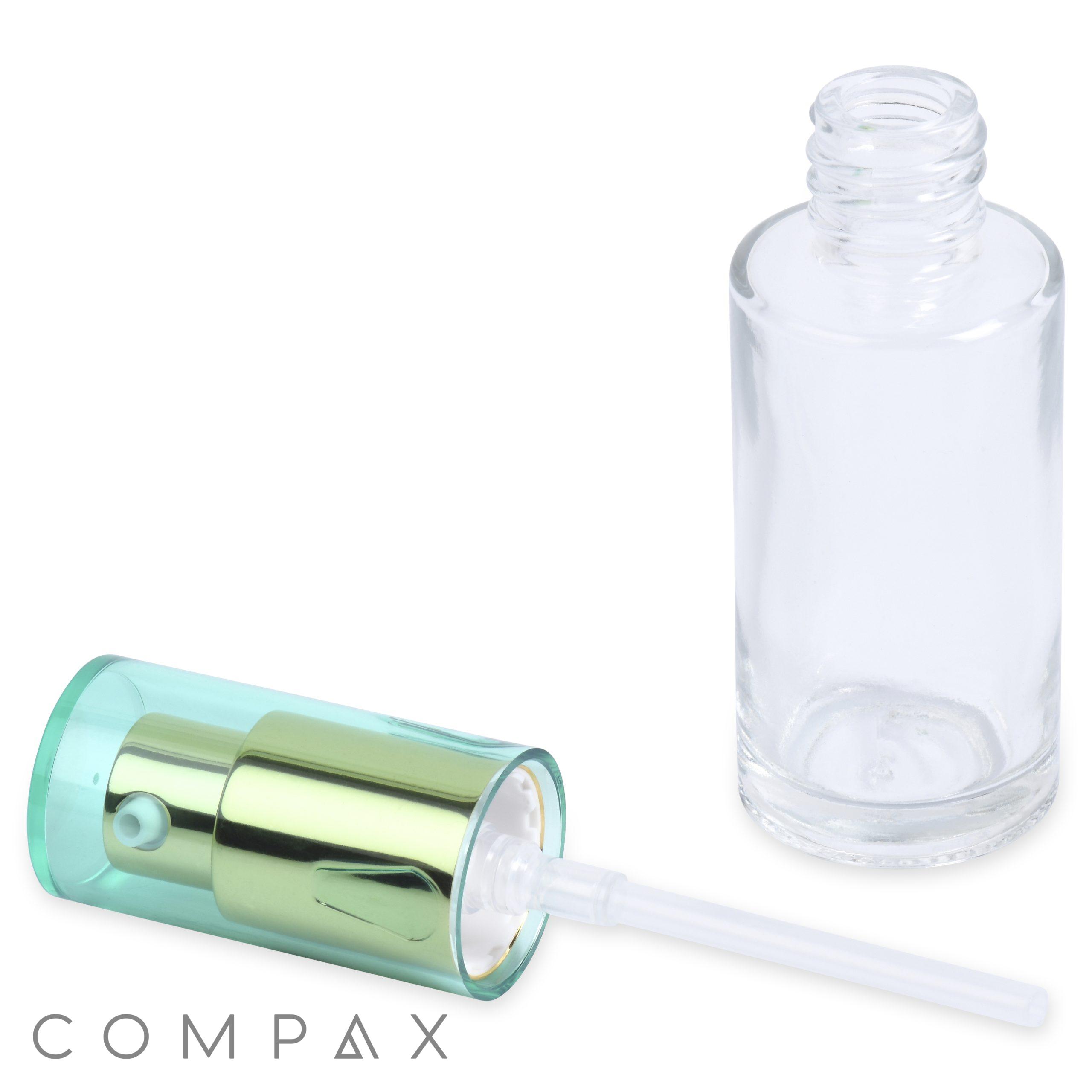 A small, clear 28mL Glass Cylinder Round Bottle lies on its side next to its detached spray cap with a metallic green and blue finish. The cap has a white plastic tube attached. The word "COMPAX" is visible at the bottom left corner of the image.