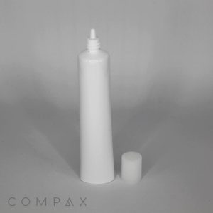 A 25mm No-Drip Airless Valve Tip Tube with a white pointed nozzle stands upright in the center. The white cap, removed from the tube, is placed next to it on the right. The background is plain and light gray. The word "COMPAX" is subtly printed at the bottom left corner.