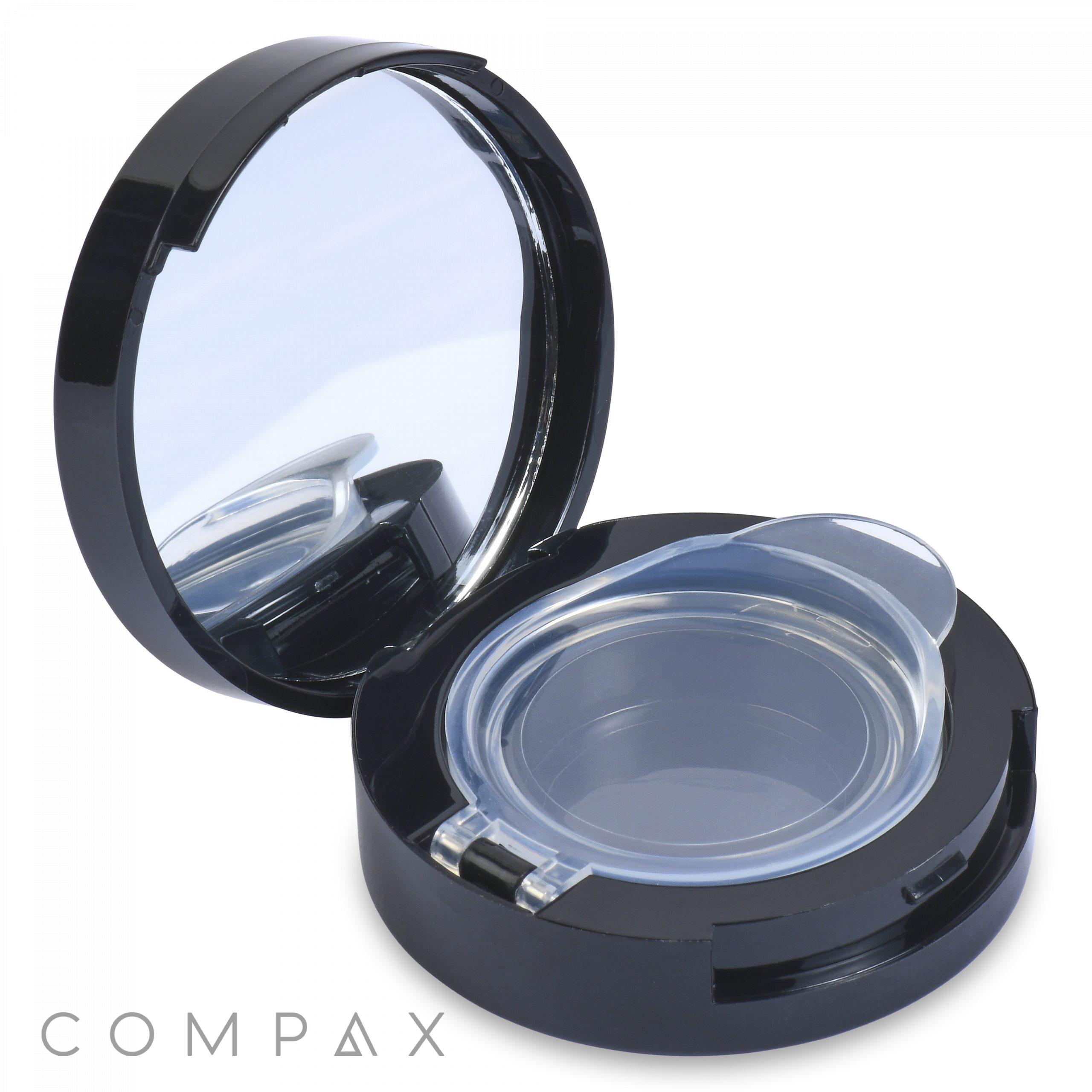 A round black compact case, the Airtight Eyeshadow Compact, is open, revealing an empty clear plastic compartment along with a built-in mirror on the inside of the lid. The word "COMPAX" is displayed in light gray at the bottom-left corner of the image.