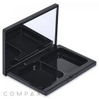 A black, square Double Pan Compact is open, revealing two empty compartments and a mirror on the interior side of the lid. One compartment features a grid pattern on its base. The word "COMPAX" is printed in grey on the white background below the compact.
