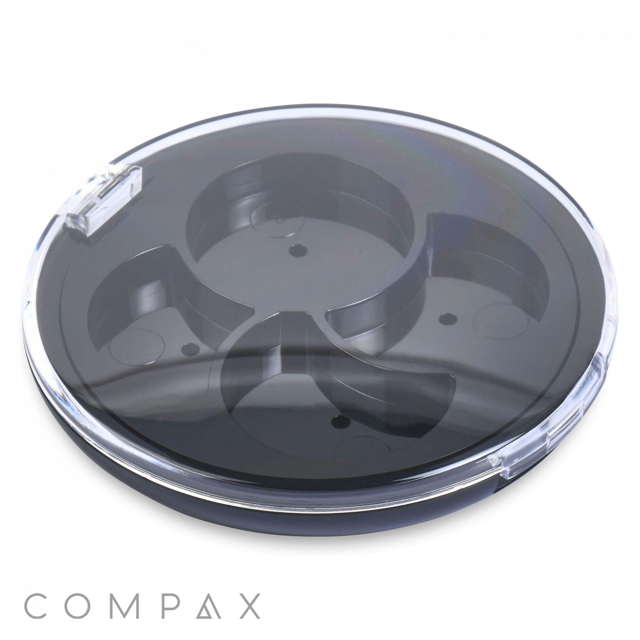 The Round 4 Pan Pinwheel Eye Shadow Compact is a transparent, circular container divided into four compartments with a snap-on lid. The base of this compact is black, and the clear lid showcases the sections inside. The word "COMPAX" can be seen in the bottom left corner on the outside of the container.