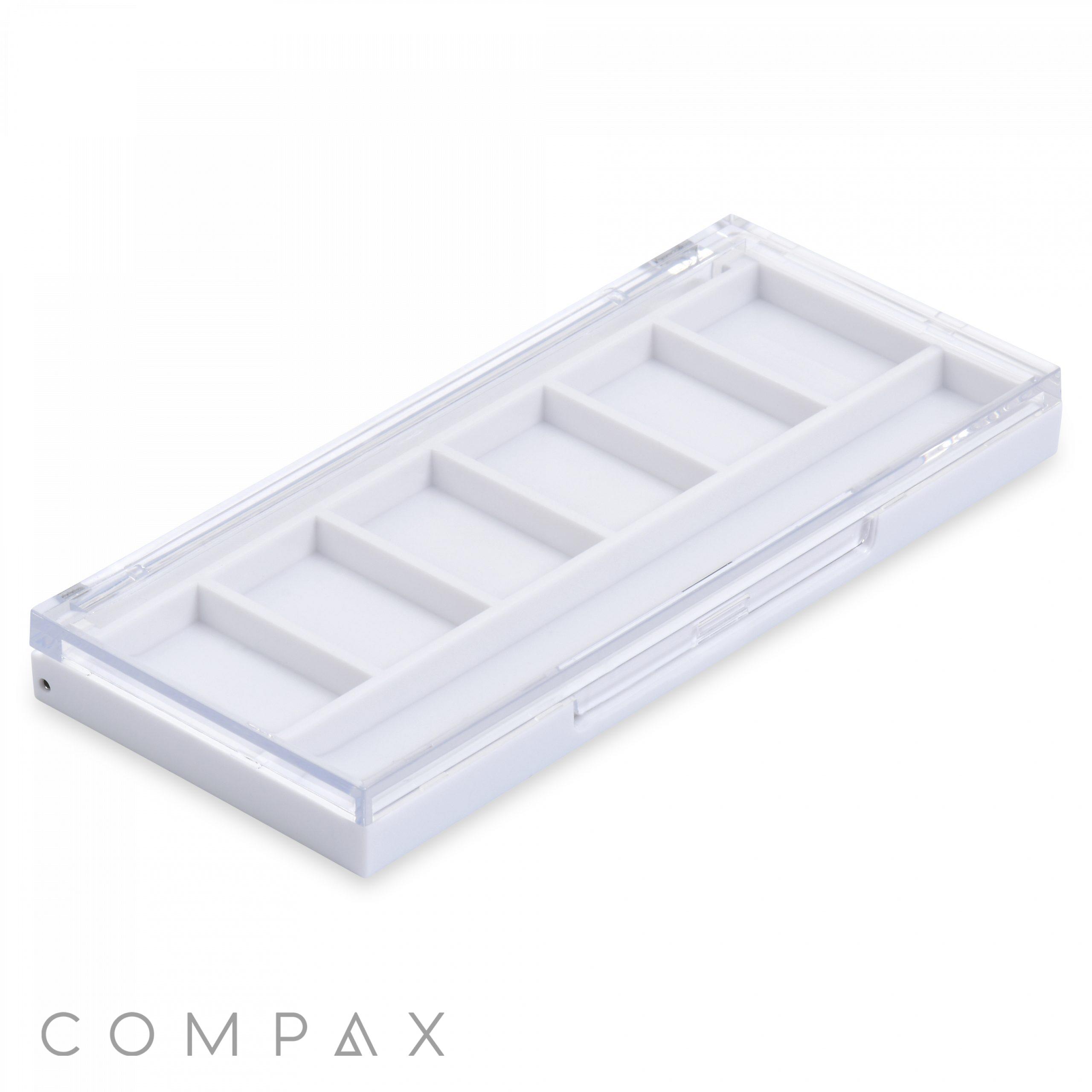 A Square Six Pan Eyeshadow Compact, featuring a white rectangular box with a clear lid and six compartments inside. The box is positioned diagonally, showcasing the depth of the compartments. The word "COMPAX" is visible in grey letters at the bottom left corner of the image.