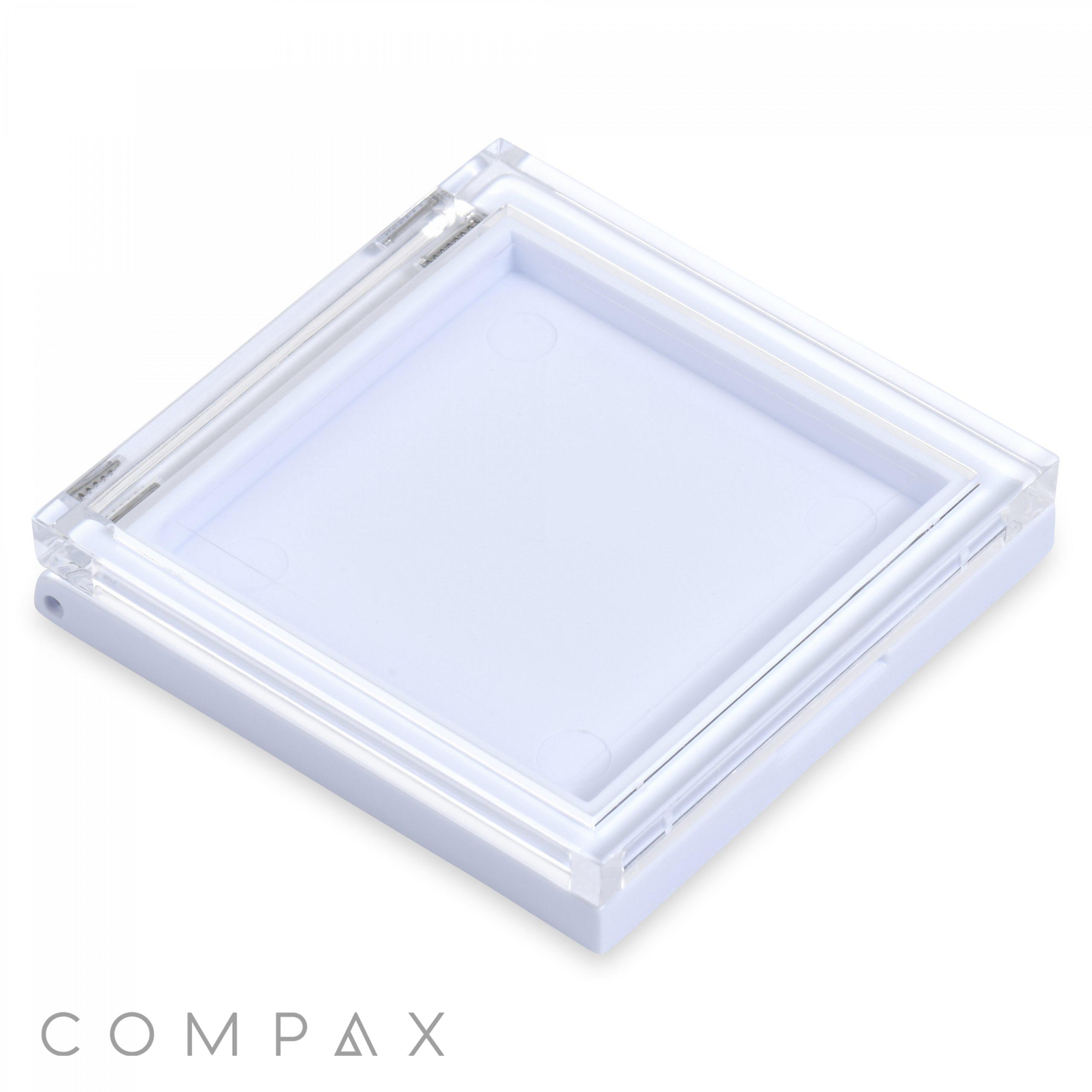 The Square Single Pan Compact features a transparent square design with a white base and a clear lid. The lid has angular corners and is slightly raised above the base. "COMPAX" is written in gray capital letters on the bottom-left corner, with a white background.