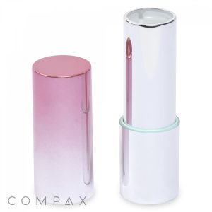 A sleek, metallic case of the Round Lipstick is showcased with its lid removed. The lid features a pink gradient design, while the base is a shiny silver accented with a mint green ring. The brand name "COMPAX" appears in the lower left corner.