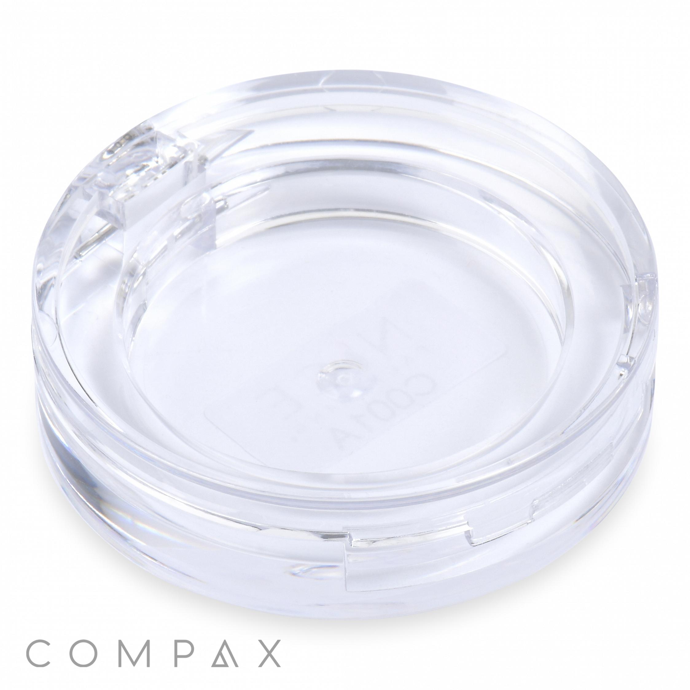 A Round Single Pan Eyeshadow Compact is displayed against a white background. The clear plastic compact case, featuring a transparent lid, appears empty. The brand name "COMPAX" is visible at the bottom left corner of the image.