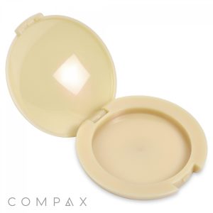 A beige Round Single Pan Compact is open, displaying a matching interior where the powder can be stored. The case features a hinge at the back and a latch at the front. The word "COMPAX" is printed on the bottom left corner of the exterior.
