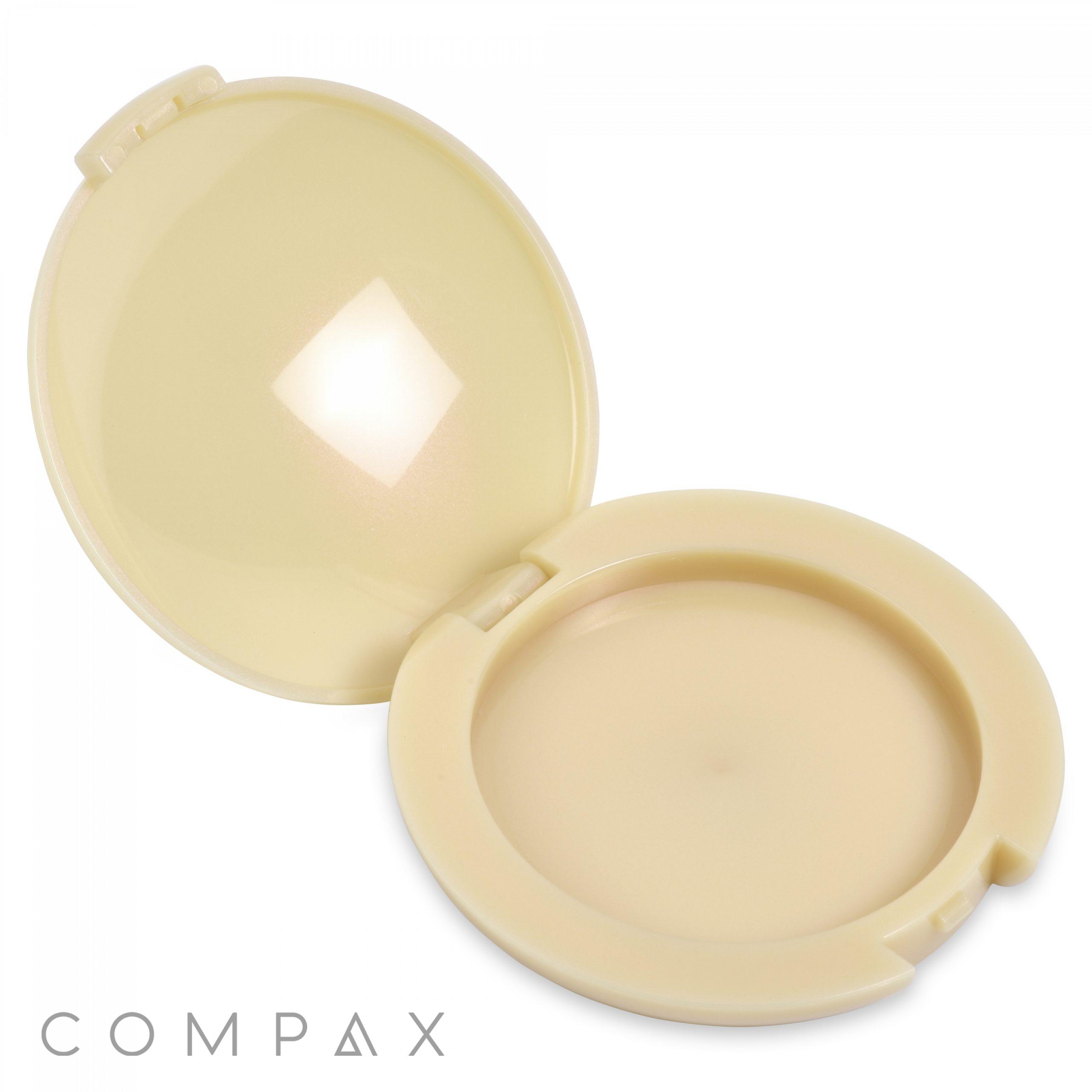 A beige Round Single Pan Compact is open, displaying a matching interior where the powder can be stored. The case features a hinge at the back and a latch at the front. The word "COMPAX" is printed on the bottom left corner of the exterior.