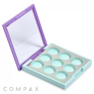 A square, open makeup palette with a mint green base and nine circular compartments for storing makeup. It features a built-in mirror on the inside of the lavender-colored lid. The brand name "COMPAX" is visible in the lower left corner.
