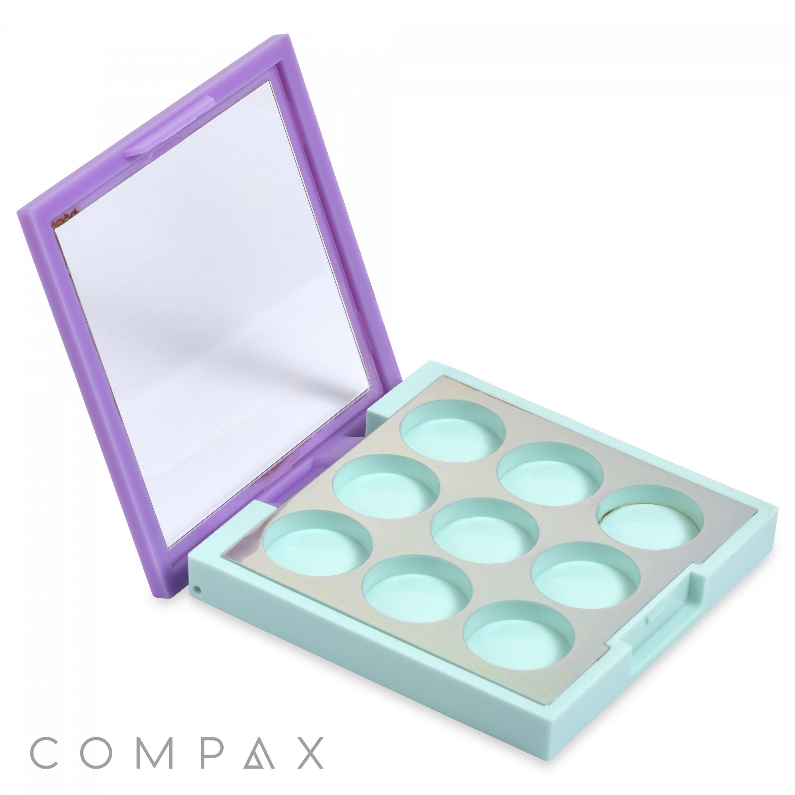 A square, open makeup palette with a mint green base and nine circular compartments for storing makeup. It features a built-in mirror on the inside of the lavender-colored lid. The brand name "COMPAX" is visible in the lower left corner.