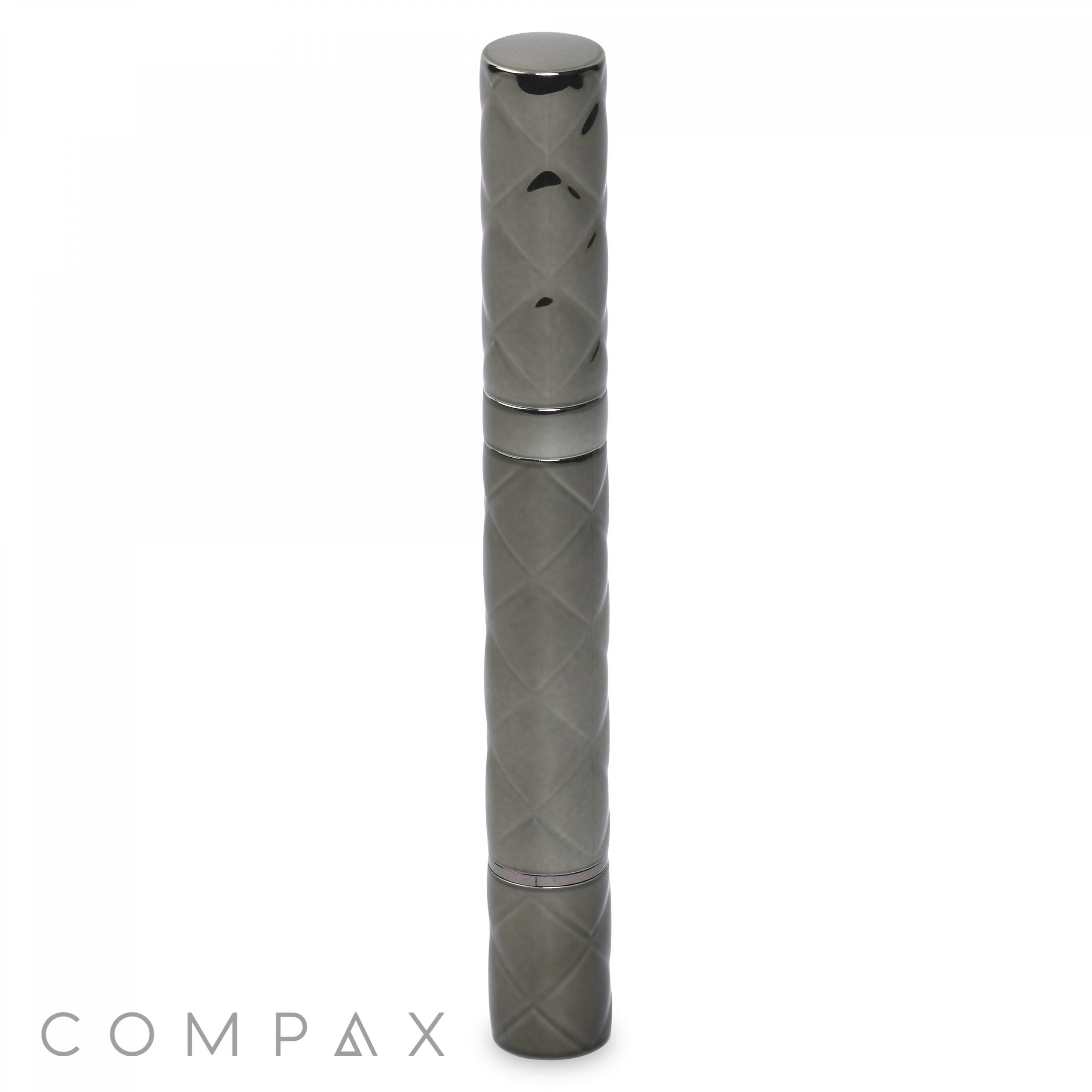 A sleek gray cylindrical object with a quilted pattern, likely the Round Lipstick, stands vertically against a white background. The word "COMPAQ" is displayed at the bottom left corner in a modern font.