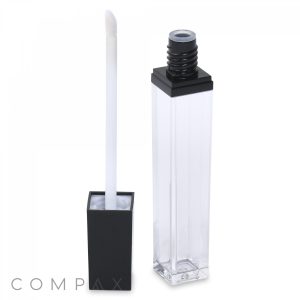 A transparent, square lip gloss container with a black cap is shown. The cap is removed, revealing an attached white applicator wand resting next to the open container. The brand name "COMPAX" is subtly positioned in the lower left corner of the container.