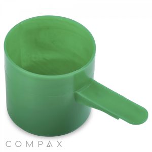 A 67cc green plastic measuring scoop with a short handle is displayed empty against a white background. The word "Compax" appears in light gray text at the bottom of the image.