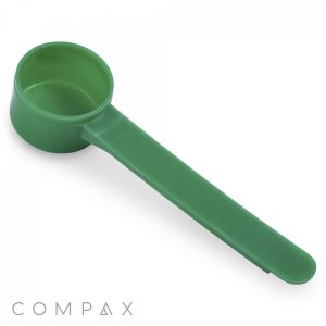 An 11cc green plastic measuring scoop with a long handle, lying on a white surface. The scoop has a cylindrical measuring cavity on one end and a flat, narrow handle extending from it. The brand name "COMPAX" is visible at the bottom left corner.