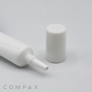 A 16mm Needle Nose white plastic squeeze tube with a small nozzle, likely for ointment or cream, lies on its side. The tube's cap is standing upright next to it, detached from the nozzle. The background is plain white, and "COMPAX" is written in the bottom left corner.
