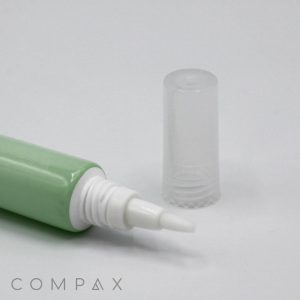 A close-up image displays a green tube labeled "19mm Brush Applicator" with a white applicator tip. The clear, cylindrical cap has been removed and placed next to the tube, revealing the fine tip. The brand name "COMPAX" is visible at the bottom corner of the image.