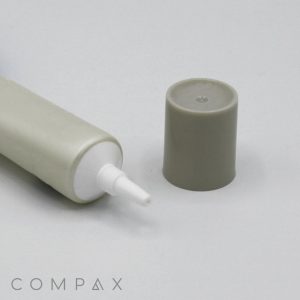 A white tube labeled "19mm Needle Nose" featuring a grey nozzle and a matching grey cap is arranged horizontally on a flat surface. The cap lies adjacent to the tube. The word "COMPAX" appears in the bottom left corner of the image, complementing the minimalistic and neutral color scheme.