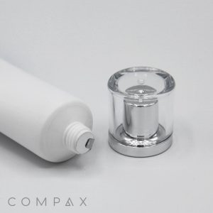 The image depicts a white cylindrical cosmetic bottle lying on its side, with its clear, silver-colored 25mm Screw Top cap removed and placed next to it. The cap features a transparent outer layer and a reflective interior. The word "COMPAX" is visible in light gray text in the bottom left corner.