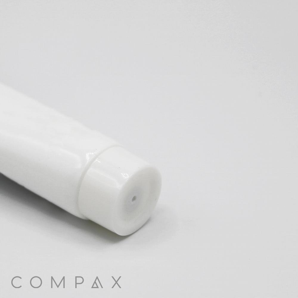 A 25mm Screw Top, white and cylindrical in shape, is laying horizontally on a light gray surface. The focus is on its capped end, with the word "COMPAX" appearing in gray text at the bottom left corner of the image.