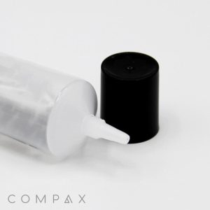 A close-up image of a transparent plastic bottle lying horizontally with a thin, 25mm Needle Nose tip exposed. The black bottle cap is positioned beside the nozzle. The background is a clean, white surface. The word "COMPAX" is visible in the bottom left corner.