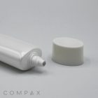A white tube with a nozzle lies horizontally on a grey surface, its cap removed and placed nearby. The brand name "COMPAX" is printed in small, grey text at the bottom left corner of the image. The product is named 30mm Flat Screw Top.