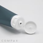 A close-up of the 35mm Flip Top, a gray tube with a white flip-top cap, partially open, lying sideways on a white surface. The word "COMPAX" is visible in simple font at the lower left corner of the image.