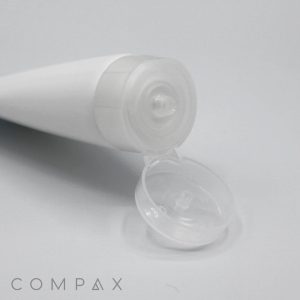 A close-up image of an open white squeeze tube with a clear 38mm flip-top cap. The tube is placed on a light surface with the cap lying directly in front of the tube's opening, showing the nozzle. The word "COMPAX" is printed in the bottom left corner.
