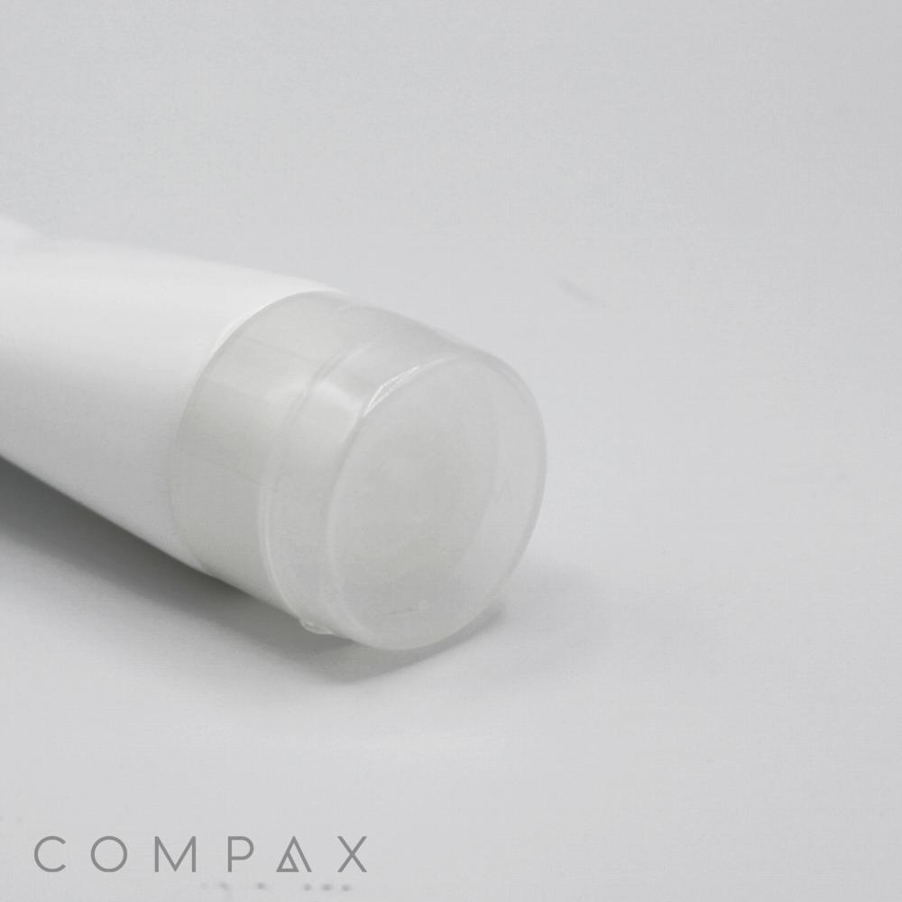 An up-close image of a white tube with a transparent cap, placed on a plain white background. The brand name "COMPAX" is visible in the bottom left corner. The tube, labeled as "38mm Flip Top," appears to be a cosmetic or skincare product.