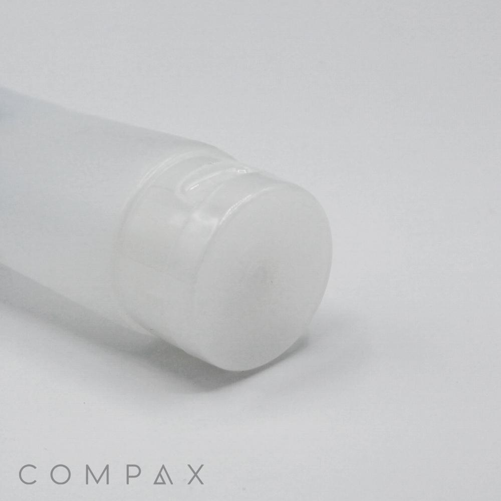 A close-up of a semi-transparent, cylindrical plastic bottle with a white cap lying horizontally on a white surface. The cosmetic bottle, named "40mm Flip Top," features minimal branding with "COMPAX" written in light gray text at the bottom left corner.