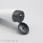 A close-up of a white cylindrical bottle with a gray flip-top cap is shown. The cap is open, and the bottle lies on its side, highlighting the opened top. The word "COMPAX" can be seen at the bottom left corner of the image.