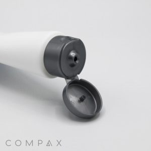 A close-up of a white cylindrical bottle with a gray flip-top cap is shown. The cap is open, and the bottle lies on its side, highlighting the opened top. The word "COMPAX" can be seen at the bottom left corner of the image.