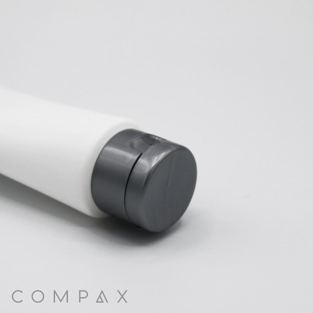 A white cylindrical tube labeled "40mm Flip Top," featuring a gray flip-top cap, is positioned horizontally on a white surface. The word "COMPAX" is printed in gray text near the bottom left corner.