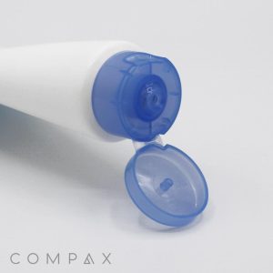 A white tube labeled "40mm Flip Top" with a blue flip-top cap, partially open to reveal the nozzle, displays a simple and minimalistic design. The word "COMPAX" appears in the bottom left corner. The tube is positioned diagonally against a plain white background.