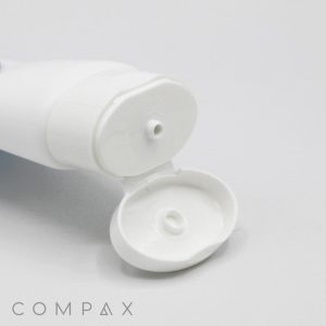 A 40mm Oval Flip Top white plastic tube with a flip-open cap lies on a plain white background. The opening of the tube is visible, showing a small nozzle for dispensing the product. The word "COMPAX" is printed near the bottom left corner of the image.