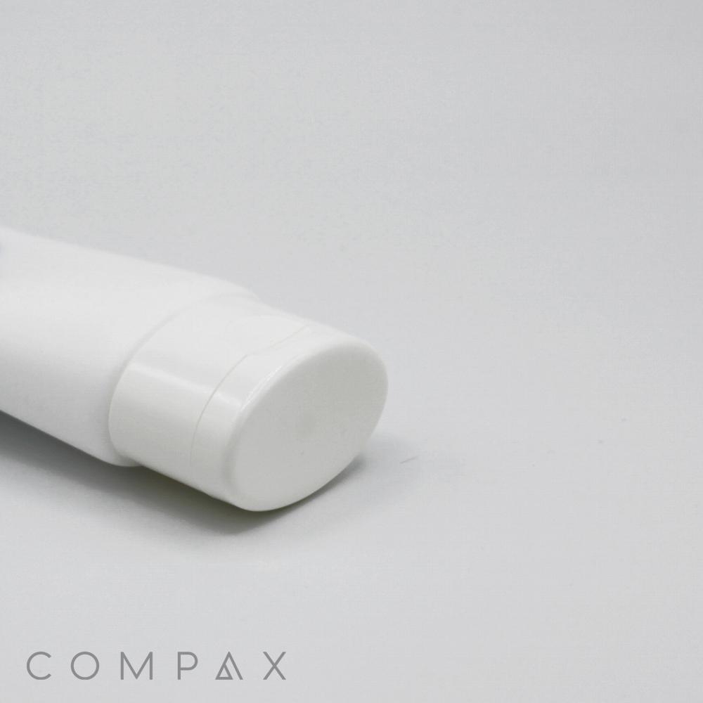 A portion of a white, cylindrical tube container with a round, flat cap is partially visible against a plain, off-white background. The word "COMPAX" is printed in light gray in the bottom left corner of the image.