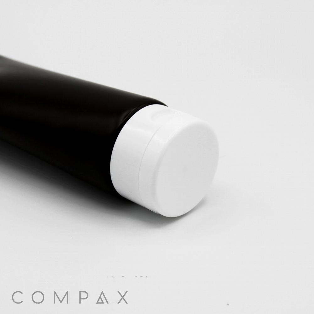A close-up of a cylindrical black tube featuring a white flip-top cap. The tube is slightly angled, and the focus is on the cap. The word "COMPAX" is visible in the bottom left corner of the image. The background is plain white.
