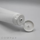 A white plastic tube from the "45mm Flip Top" product line lies on a flat surface with its flip-top cap open, revealing a small opening for dispensing contents. Both the tube and cap are white, and the plain tube surface has no visible labeling except for the word "COMPAX" written in grey in the bottom left corner.