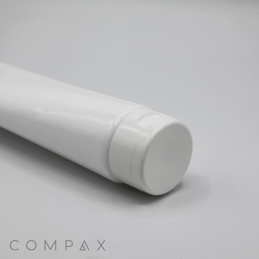 A white, cylindrical tube with a flip top lies on a smooth, light-colored surface. The tube appears to be unused and clean, suggesting it's for a cosmetic or personal care product. The word "COMPAX" is subtly printed in grey text in the bottom left corner.