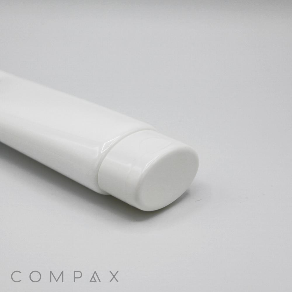 A close-up shot of a white, oval tube with a flip-top cap placed against a plain white background. The word "COMPAX" is printed at the bottom left corner in a minimalist font.