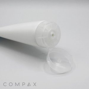 A white cylindrical tube with a clear flip-top cap lies on a flat surface, open and facing upwards. The product name "50mm Flip Top" and the brand name "COMPAX" are visible in the lower left corner. The background is plain and light grey.