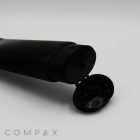 A 50mm Oval Flip Top squeeze tube in black, lying on its side with the flip-top cap open. The tube is set against a plain white background, with the word "COMPAX" in light gray letters partially visible at the bottom left corner.