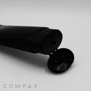 A 50mm Oval Flip Top squeeze tube in black, lying on its side with the flip-top cap open. The tube is set against a plain white background, with the word "COMPAX" in light gray letters partially visible at the bottom left corner.
