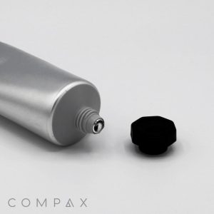 A 40mm Screw Top tube in metallic gray lies on a white surface with its black cap removed and placed beside it. The tube's opening is visible, and the text "COMPAX" is printed in the bottom left corner.