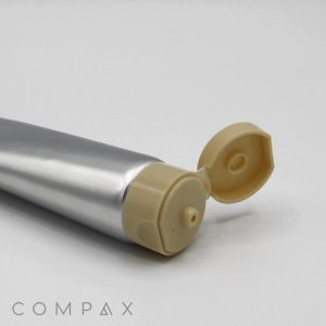 A silver-colored tube, named "35mm Flip Top," with a beige flip-top cap lies on a white surface. The cap is open, revealing the nozzle from which the tube's contents can be dispensed. The word "COMPAX" is visible in the bottom left corner of the image.