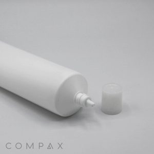 A white 40mm Needle Nose cylindrical tube with its cap removed, laying horizontally on a light grey surface. The tube's dispensing nozzle is visible, and the cap is placed beside it. The word "COMPAX" is subtly printed in the bottom left corner.