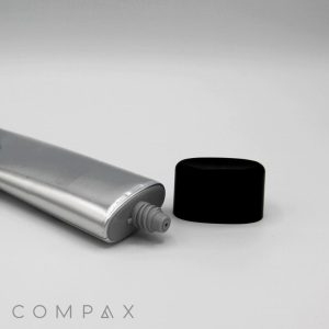 A 40mm Flat Screw Top in silver, with its black cap removed, rests on a white surface. The cap is placed to the right of the tube, and the brand name "COMPAX" is visible in small letters at the bottom left corner of the image.